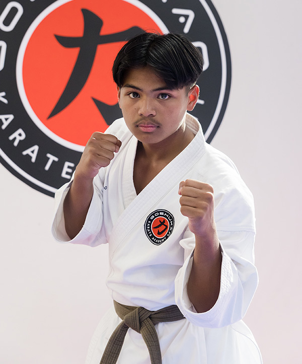 Biensy Knight in a fighting pose in front of the large Goshukan Karate Academy club badge at Seven Hills dojo.